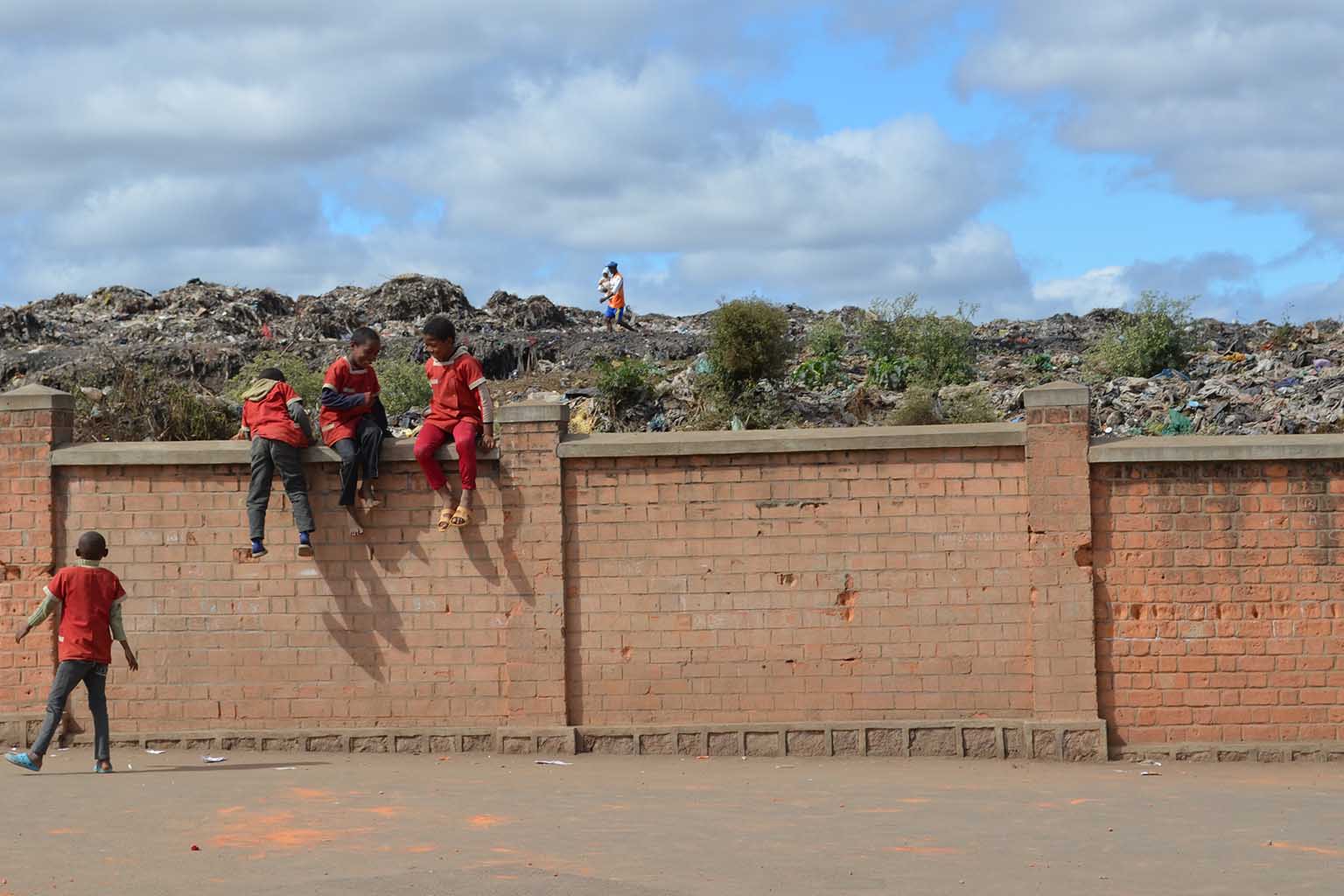 Children sit on a brick wall outside of a garbage heap.